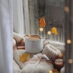 Warm autumn setting with coffee, sweater, and leaves by rainy window.