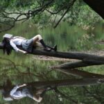 A young woman with a book on her face lies relaxing on a tree trunk by a tranquil lake.